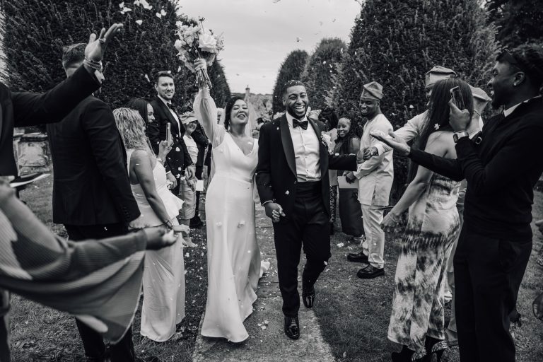 Elmore Court wedding photography, bride and groom smile and raise flowers as they walk through guests