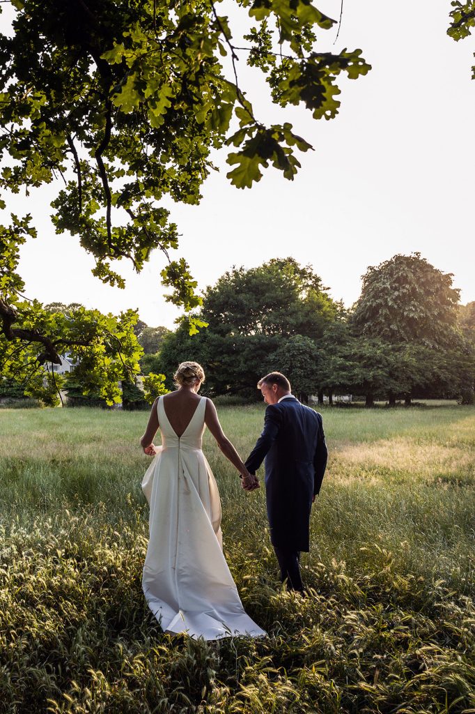 Bride and groom holding hands as they walk through a long grassed field under a tree illuminated by evening sunlight in South Wales